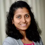 Smiling woman with long, wavy black hair, wearing a gray cardigan over a pink top, stands against a blurred, neutral background. She has a small red bindi on her forehead.