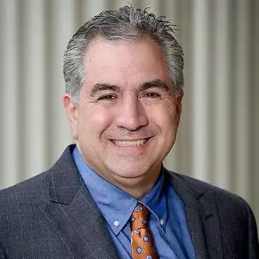 A smiling man with short gray hair is wearing a dark gray suit, blue shirt, and orange patterned tie. The background features a series of vertical lines in soft focus.