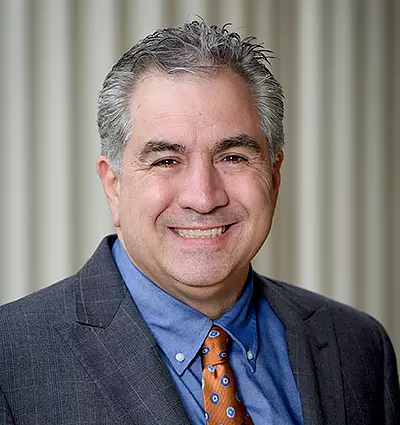 A smiling man with short gray hair is wearing a dark gray suit, blue shirt, and orange patterned tie. The background features a series of vertical lines in soft focus.