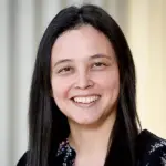 A person with long black hair is smiling, wearing a black top with floral patterns. The background is softly blurred in neutral tones.