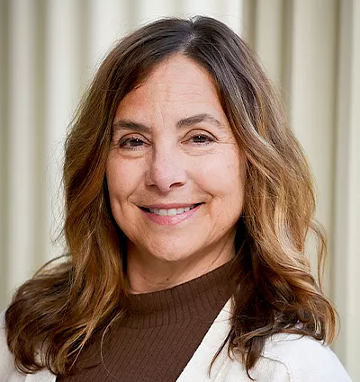 A woman with long, wavy brown hair smiles while wearing a white sweater over a brown turtleneck. The background features vertical cream-colored blinds.