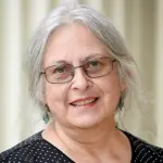 A woman with long gray hair and glasses smiles at the camera. She is wearing a black polka dot top and teal earrings, standing in front of a light-colored, blurred background.