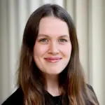 Portrait of a smiling person with long brown hair, wearing a black top. They are standing in front of a light-colored, softly blurred background.
