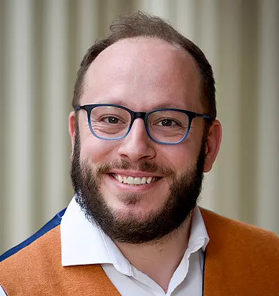 Smiling man with glasses and a beard, wearing a white shirt and an orange vest, standing in front of a blurred background.