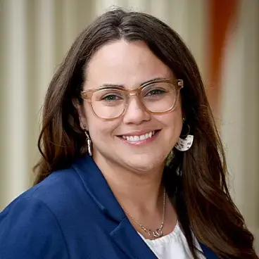 A person with long brown hair and glasses is smiling at the camera. They are wearing a blue blazer, a light-colored blouse, and drop earrings. The background is softly blurred, suggesting an outdoor setting.