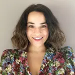 A person with shoulder-length wavy hair smiles at the camera. They are wearing a colorful floral top with a neutral background behind them.