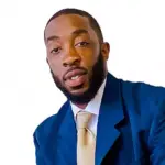 A man with a beard wearing a blue suit and a light-colored tie against a plain white background. He is looking toward the camera.