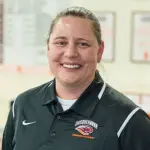 A person wearing a black polo shirt with a logo and text that says "Susquehanna Swimming & Diving" stands indoors, smiling. The background is blurred, showing some wall decorations.