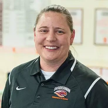 A person wearing a black polo shirt with a logo and text that says "Susquehanna Swimming & Diving" stands indoors, smiling. The background is blurred, showing some wall decorations.