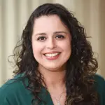 A woman with long, curly dark hair smiles warmly at the camera. She is wearing a dark green top and a delicate necklace with a pendant. The background is softly blurred with neutral tones.