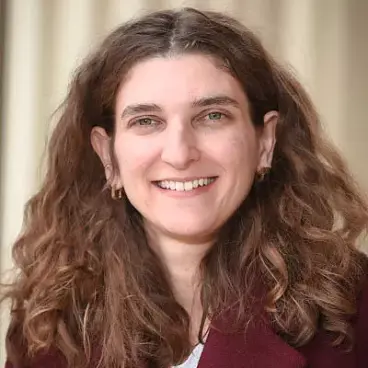 Woman with long, wavy brown hair, smiling. She's wearing a burgundy jacket and a light-colored top. The background is softly blurred with neutral tones.