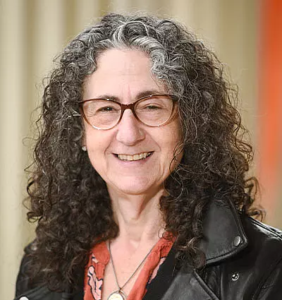 A person with curly gray hair and glasses smiles at the camera. They are wearing a black jacket over a coral shirt with a floral pattern. The background is softly blurred.