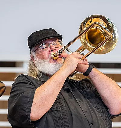 A bearded man wearing glasses and a black cap plays a brass trombone. He is dressed in a dark shirt and stands in front of a blurred background, concentrating on his performance.