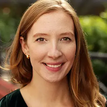 A woman with long reddish hair smiles warmly while looking at the camera. She is outdoors with a blurred background of greenery. She is wearing gold hoop earrings and a dark top.