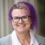 Person with curly purple hair and glasses, wearing a gray blazer over a light pink shirt, smiles at the camera. The background is blurred, with vertical lines suggesting a structure.