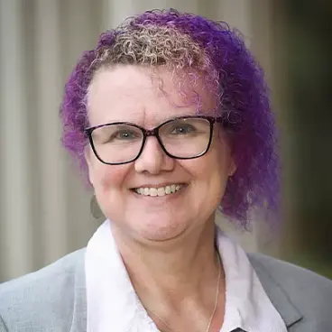 Person with curly purple hair and glasses, wearing a gray blazer over a light pink shirt, smiles at the camera. The background is blurred, with vertical lines suggesting a structure.