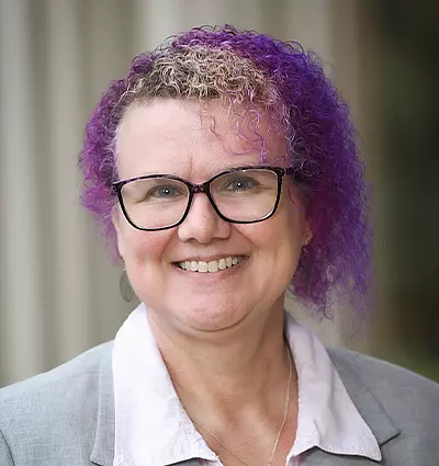 Person with curly purple hair and glasses, wearing a gray blazer over a light pink shirt, smiles at the camera. The background is blurred, with vertical lines suggesting a structure.