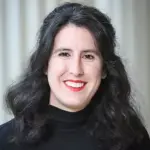 A person with long, wavy black hair and red lipstick is smiling at the camera. They are wearing a black turtleneck sweater. The background is softly blurred with vertical lines.