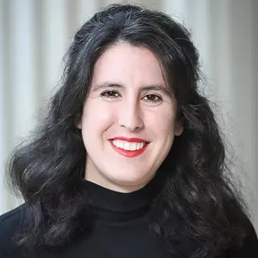 A person with long, wavy black hair and red lipstick is smiling at the camera. They are wearing a black turtleneck sweater. The background is softly blurred with vertical lines.