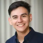 A person with short dark hair smiles warmly, wearing a dark collared shirt against a light, blurred background.