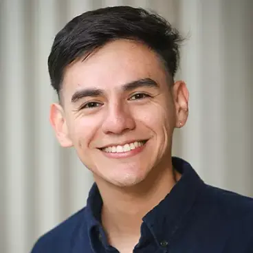 A person with short dark hair smiles warmly, wearing a dark collared shirt against a light, blurred background.