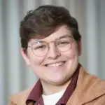 A person with short hair and glasses smiles warmly. They are wearing a brown jacket over a burgundy shirt. The background is softly blurred with vertical lines.