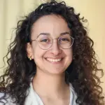 Smiling person with curly hair, wearing glasses and a white shirt, stands in front of a neutral background.