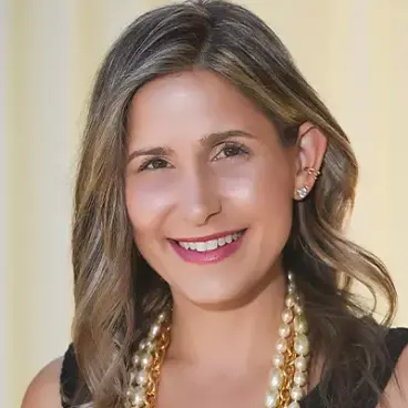 A woman with wavy, shoulder-length hair smiles at the camera. She is wearing a black top and a double-layered pearl necklace, against a soft, light background.