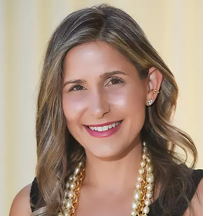 A woman with wavy, shoulder-length hair smiles at the camera. She is wearing a black top and a double-layered pearl necklace, against a soft, light background.