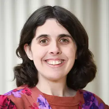 A person with shoulder-length dark hair smiling, wearing a floral-patterned blouse. The background features vertical light-colored panels.
