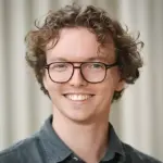 Man with curly hair, glasses, and a green shirt smiles in front of a neutral background.