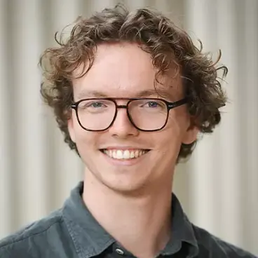 Man with curly hair, glasses, and a green shirt smiles in front of a neutral background.