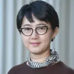 A person with short black hair and glasses smiles softly at the camera. They are wearing a dark top and a black-and-white patterned scarf around their neck, standing against a neutral, blurred background.
