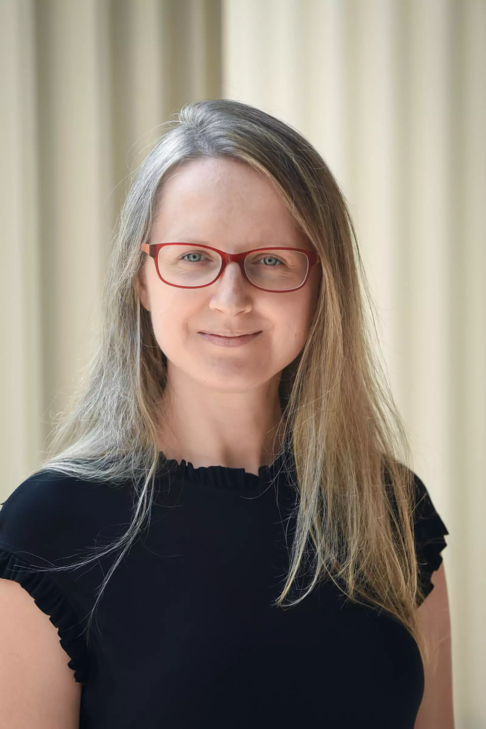 A person with long blonde hair and red glasses is smiling softly while wearing a black top. The background is blurred vertical beige stripes.