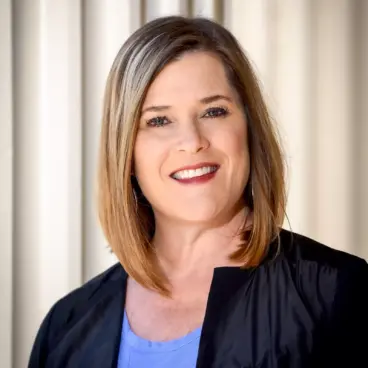 A person with shoulder-length brown hair and a bright smile is wearing a blue top and a dark jacket. They are standing in front of a light-colored, vertical-striped background.