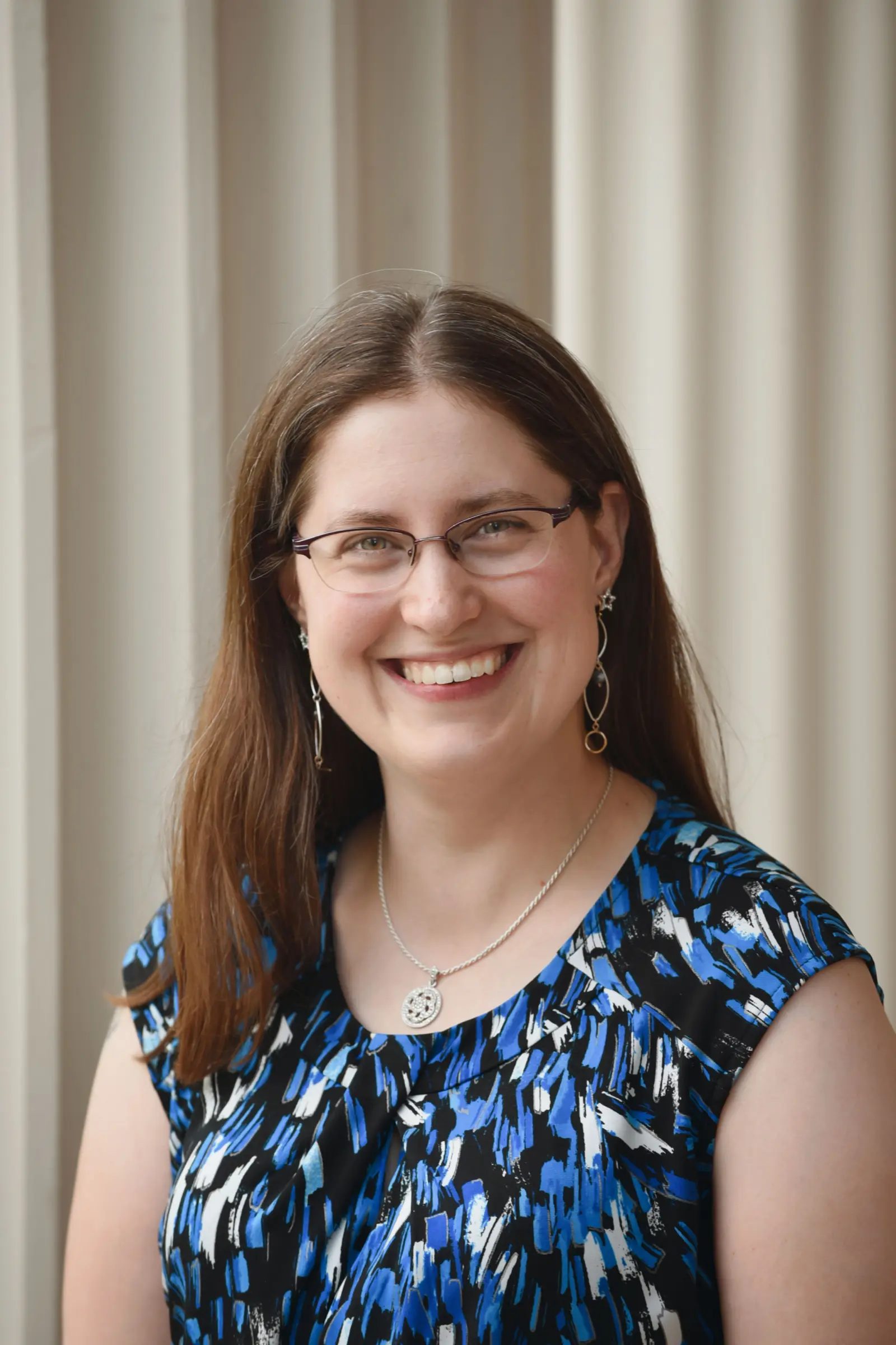 A woman with long brown hair and glasses smiles warmly. She is wearing a blue and black patterned top and a necklace with a circular pendant. The backdrop features neutral-colored vertical panels.