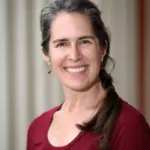 A woman with long dark hair, partially gray, smiles at the camera. She is wearing a long-sleeve maroon shirt and small earrings. The background is blurred with soft vertical lines.