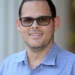 A man wearing black glasses smiles at the camera. He has short dark hair and is dressed in a blue and white checkered shirt. The background is softly blurred, with vertical lines suggesting pillars or columns.