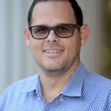 A man wearing black glasses smiles at the camera. He has short dark hair and is dressed in a blue and white checkered shirt. The background is softly blurred, with vertical lines suggesting pillars or columns.