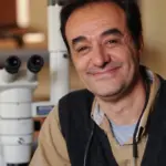 A person smiling at the camera, sitting next to a microscope in a laboratory setting. They are wearing a dark vest over a beige shirt and have reading glasses hanging from their neck.