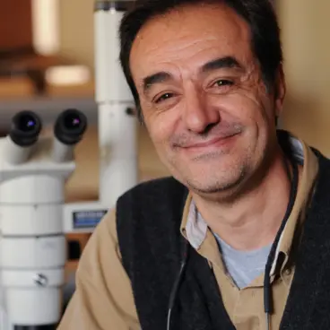 A person smiling at the camera, sitting next to a microscope in a laboratory setting. They are wearing a dark vest over a beige shirt and have reading glasses hanging from their neck.