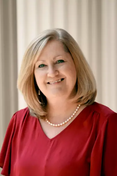 A woman with shoulder-length blonde hair smiling, wearing a red blouse and a pearl necklace. The background is softly blurred, highlighting her friendly expression.