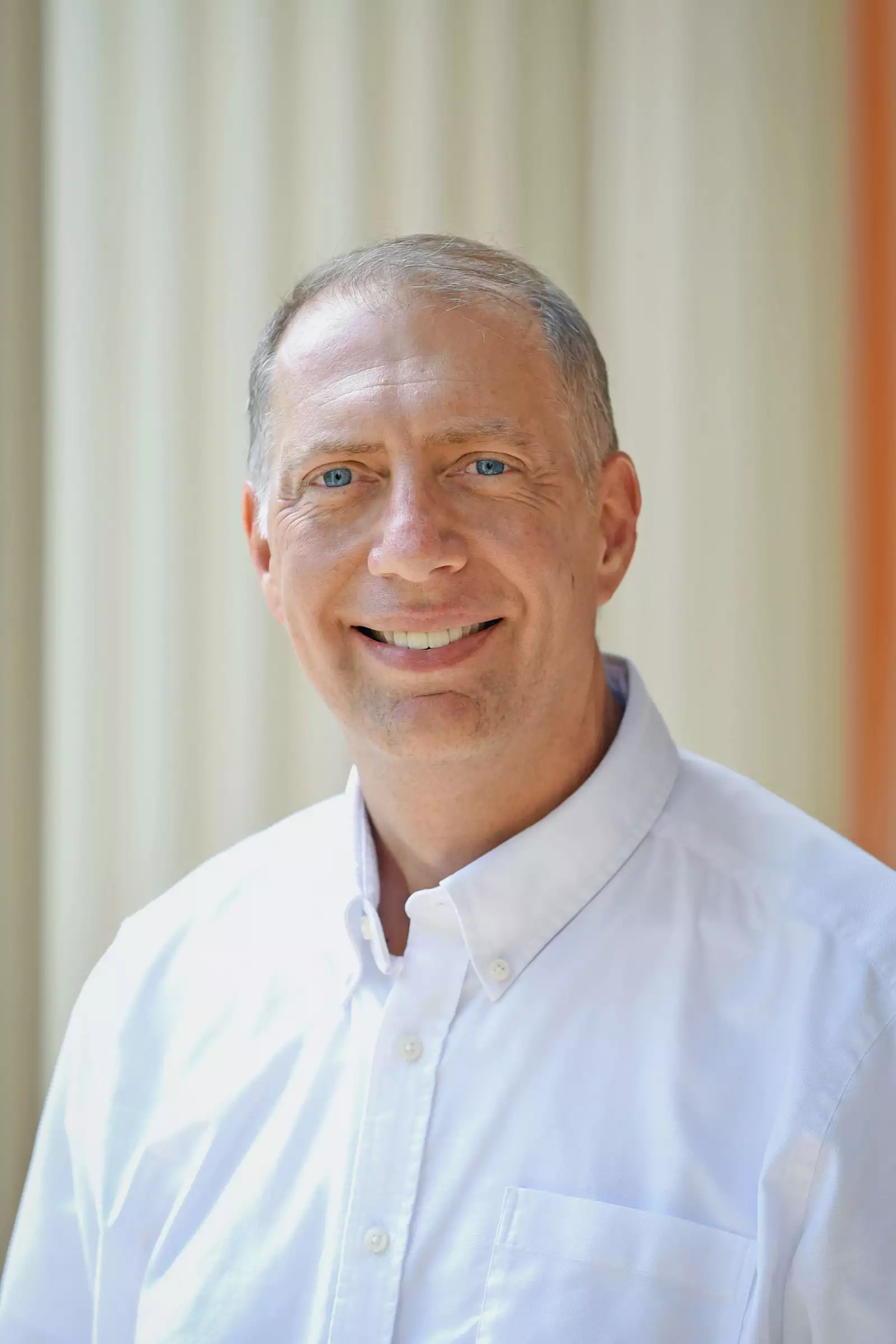 A man wearing a white button-up shirt smiles at the camera. The background features vertical, light-colored lines.
