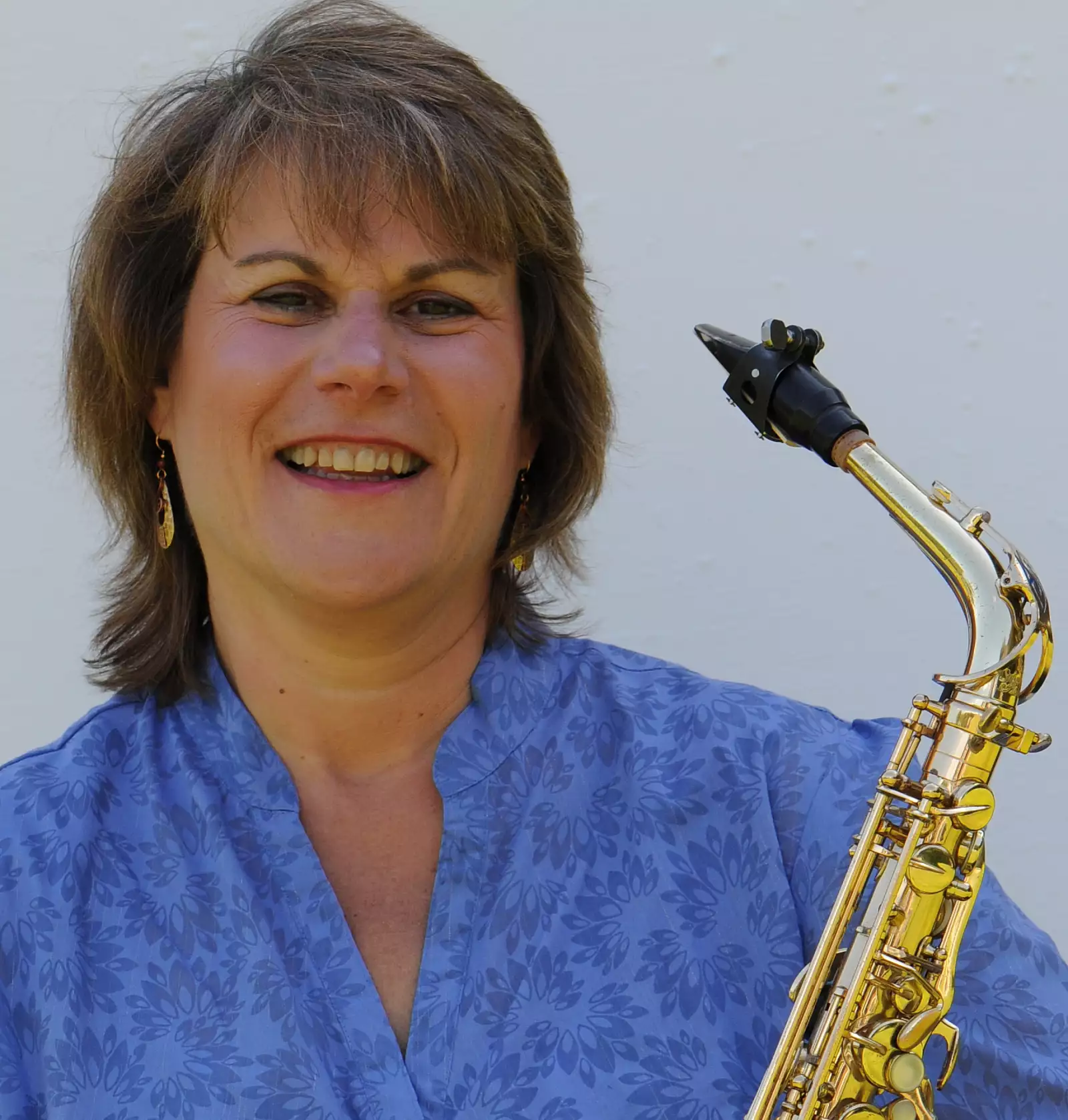 A woman wearing a blue floral blouse is smiling while holding a saxophone. The background is a plain light color.