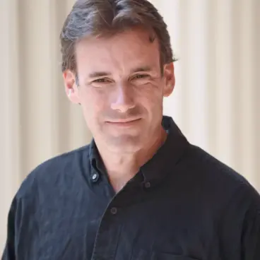 A man with short brown hair is wearing a black button-up shirt. He is standing in front of a light-colored, blurred background, smiling at the camera.