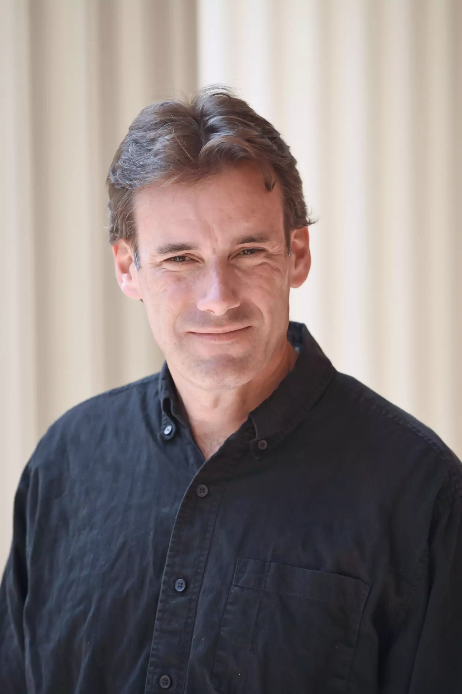 A man with short brown hair is wearing a black button-up shirt. He is standing in front of a light-colored, blurred background, smiling at the camera.