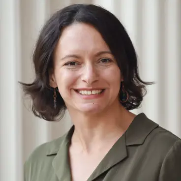 A person with short, dark hair and earrings smiling, wearing an olive green top, stands in front of a light-colored, vertically striped background.