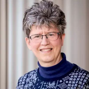 A person with short gray hair and glasses smiles at the camera. They are wearing a navy blue turtleneck with a patterned sweater. The background is softly blurred.
