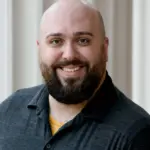 A bald man with a beard smiles at the camera. He is wearing a dark polo shirt and stands against a beige background with vertical lines.