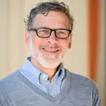 A smiling man with glasses, curly gray hair, and a beard is wearing a light blue collared shirt under a gray sweater. The background is a softly blurred indoor setting.